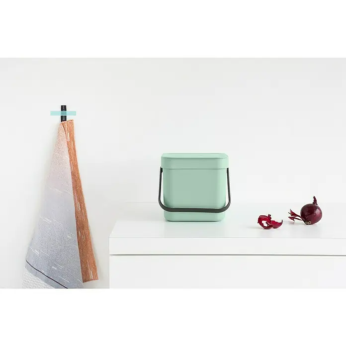 Brabantia Cubo de basura empotrado Sort & Go - 3 l, Plástico, Verde - Un cubo de compostaje verde menta sobre una superficie blanca, al lado de una cebolla roja y sus cáscaras, con un trapo de cocina al fondo.