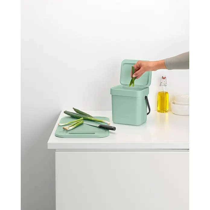 Brabantia Cubo de basura empotrado Sort & Go - 3 l, Plástico, Verde - Un cubo de basura de cocina verde menta con tapa abierta, un cuchillo y cebollas verdes sobre una tabla de cortar a juego.