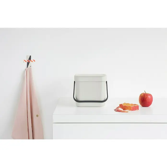 Brabantia Cubo de basura empotrado Sort & Go - 3 l, Plástico, Gris - Imagen de un elegante cubo de compost color crema sobre una superficie blanca, junto a restos de manzana y una manzana fresca, perfecto para cualquier cocina.