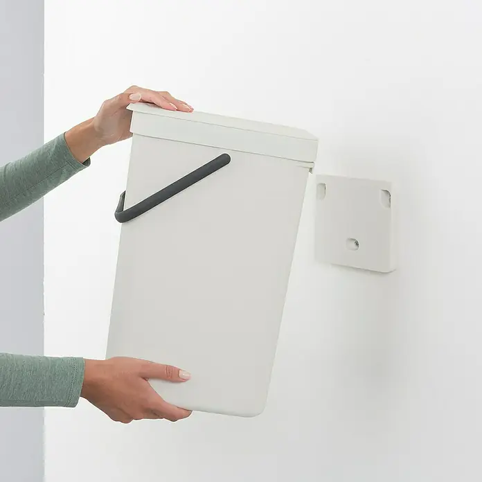 Brabantia Cubo de basura empotrado Sort & Go - 16 l, Blanco, Plástico - Un cubo rectangular blanco con asa negra, sujetado por dos manos, está a punto de ser montado en un accesorio de pared.