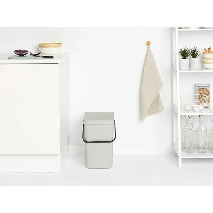 Brabantia Cubo de basura empotrado Sort & Go - 25 l, Blanco, Plástico - Un cubo de compostaje gris con asa negra se encuentra en una cocina luminosa junto a un mueble blanco y una estantería.