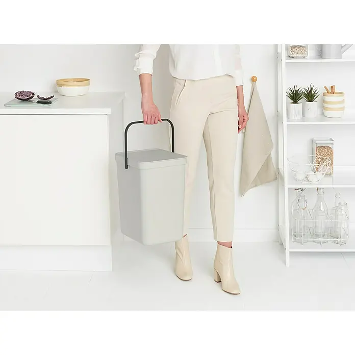 Brabantia Cubo de basura empotrado Sort & Go - 25 l, Blanco, Plástico - Una persona sostiene un cubo de basura Brabantia de color gris claro con asa negra en una cocina luminosa.