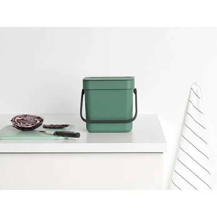 Brabantia Cubo de basura empotrado Sort & Go - 3 l, Plástico, Verde oscuro - Un contenedor de compostaje de cocina verde con asa oscura se encuentra en una encimera blanca, junto a repollo morado y un cuchillo.