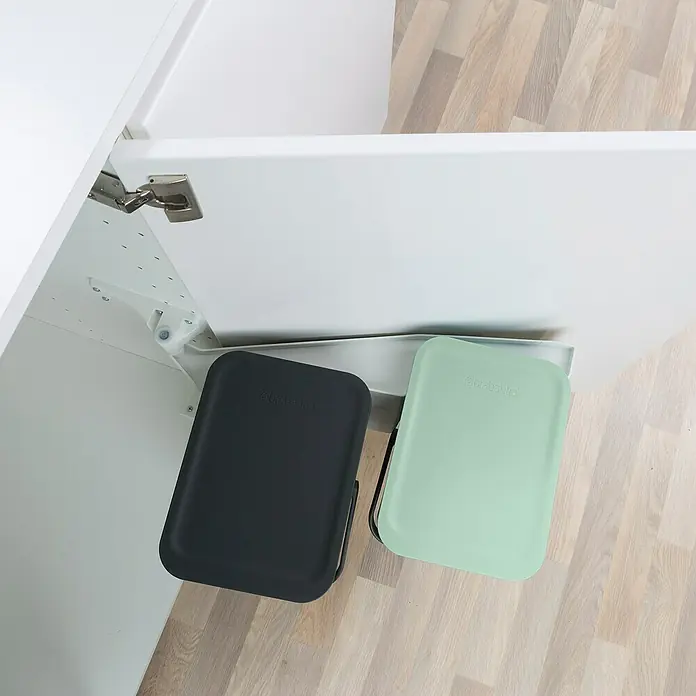 Brabantia Cubo de basura empotrado Sort & Go - 24 l, Plástico, Verde/Negro - Cubos bajo encimera: un cubo Brabanita gris y otro verde menta colgados bajo un armario blanco en una cocina con suelo de madera.