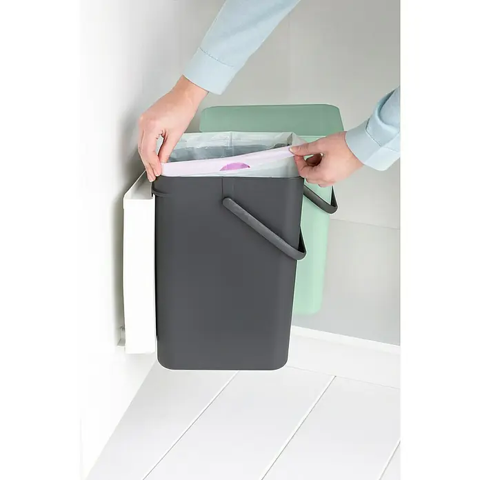 Brabantia Cubo de basura empotrado Sort & Go - 24 l, Plástico, Verde/Negro - Imagen de una persona colocando un cubo de basura con una bolsa de basura morada en un armario blanco.