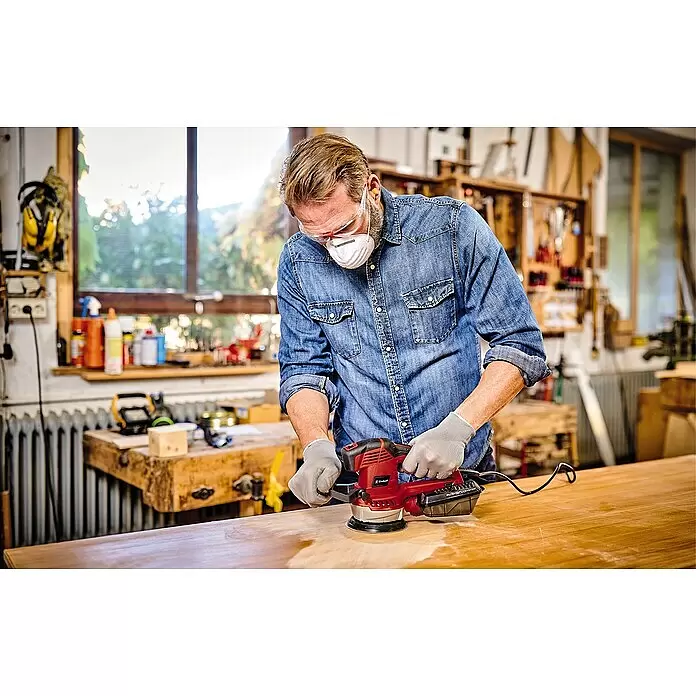 Einhell Expert Lijadora excéntrica eléctrica TE-RS 40 E - 400 W, Diámetro disco de lija: 125 mm - Un hombre usa una lijadora orbital Einhell roja para lijar una superficie de madera, con gafas de seguridad y mascarilla.