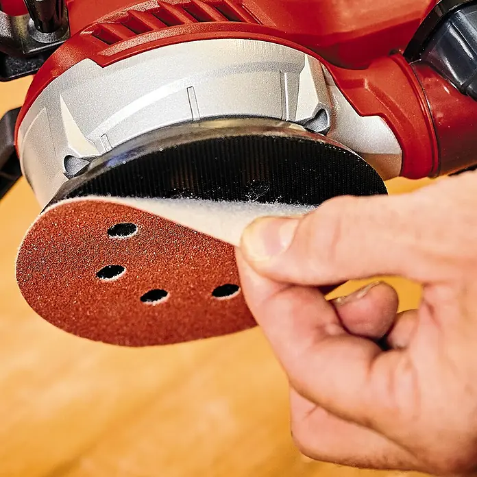 Einhell Expert Elektro-Exzenterschleifer TE-RS 40 - 400 W, Durchmesser Schleifteller: 125 mm - Nahaufnahme eines roten Exzenterschleifers mit Schleifpapier, das von einer Hand angebracht wird. Das Werkzeug ist bereit für den Einsatz.