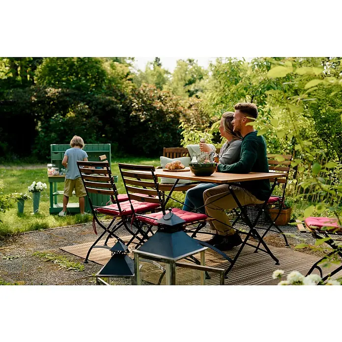 Sunfun Moni Klapptisch - L x B x H: 125 x 75 x 75 cm, Akazie, Rechteckig, Natur/Schwarz - Ein Gartentisch-Set mit Holzplatten und schwarzen Metallgestellen, ergänzt durch rote Sitzkissen, steht auf einem gewebten Teppich im Garten.