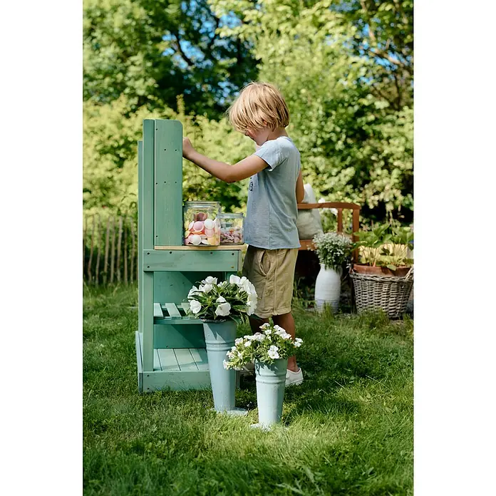 Muddy Buddy Matschküche Explorer - 102 x 40 x 105 cm, Holz, Salbeigrün - Ein Junge steht vor einem blassgrünen Verkaufsstand mit Regalen, gefüllt mit Süßigkeiten und weißen Blumen in hellblauen Vasen im Garten.