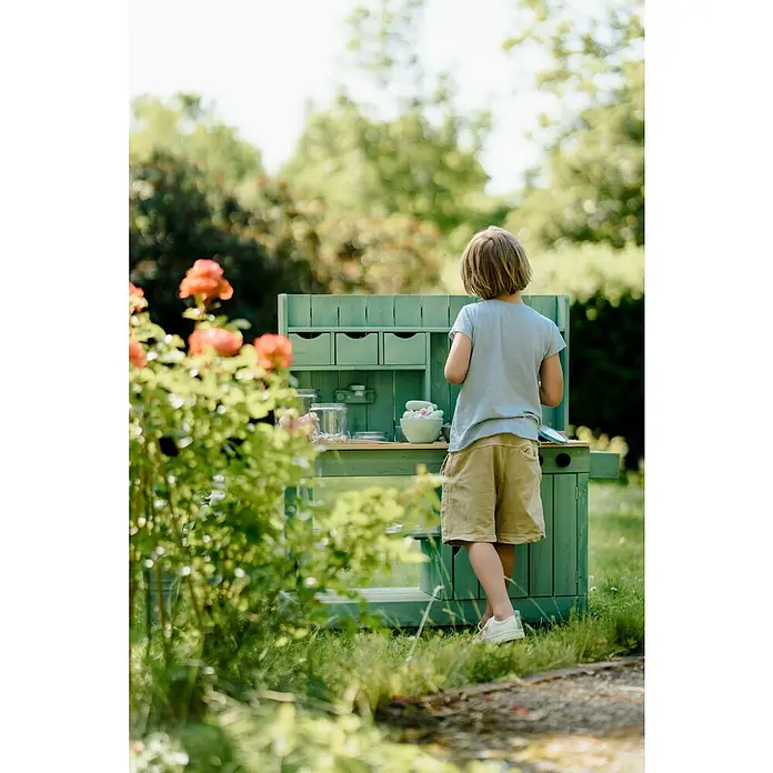 Muddy Buddy Matschküche Explorer - 102 x 40 x 105 cm, Holz, Salbeigrün - Ein Junge spielt an einem grünen Outdoor-Spielherd mit Schubladen, Gläsern und Schüsseln im Garten.