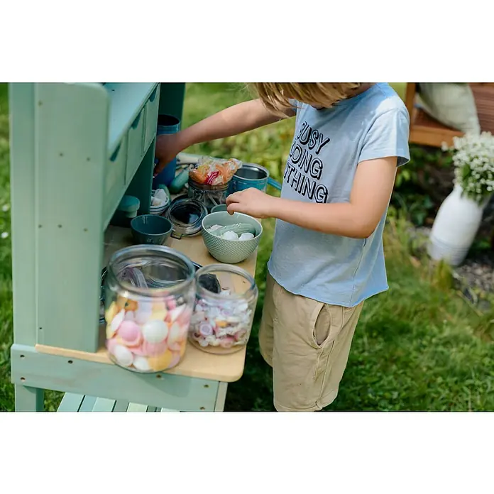 Muddy Buddy Matschküche Explorer - 102 x 40 x 105 cm, Holz, Salbeigrün - Ein Kind spielt mit einem Spielzeug-Küchenset im Freien, das mit Gläsern und Schüsseln gefüllt ist, um fantasievolles Kochen zu fördern.