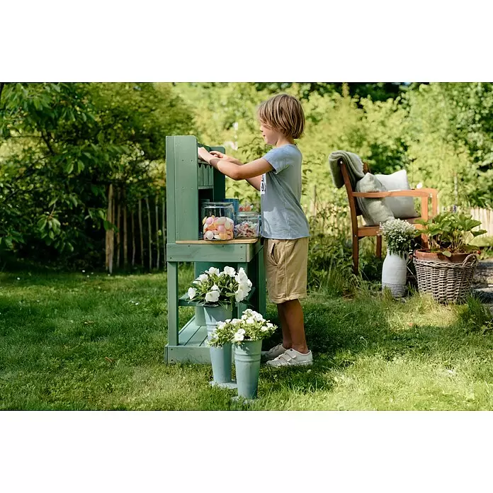 Muddy Buddy Matschküche Explorer - 102 x 40 x 105 cm, Holz, Salbeigrün - Ein Junge spielt mit einer grünen Holz-Outdoor-Spielküche mit Regalen, Bonbongläsern und Blumentöpfen in einem sonnigen Garten.