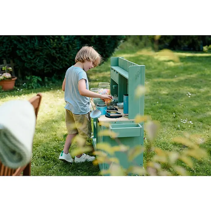 Muddy Buddy Matschküche Explorer - 102 x 40 x 105 cm, Holz, Salbeigrün - Ein Junge spielt mit einer Outdoor-Spielküche im Garten, die mit Spielzeuggeschirr und Lebensmitteln ausgestattet ist.