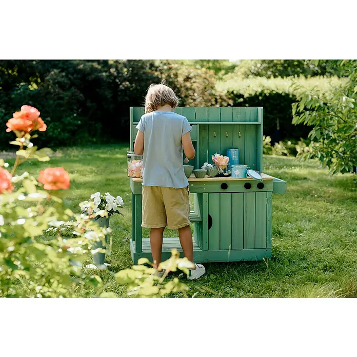 Muddy Buddy Matschküche Explorer - 102 x 40 x 105 cm, Holz, Salbeigrün - Ein Kind spielt mit einer Outdoor-Spielküche aus Holz, die mit Kochutensilien und bunten Süßigkeiten dekoriert ist, im Garten.