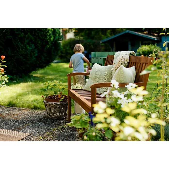 Sunfun Diana Gartenbank - B x T x H: 116 x 59 x 90 cm, Akazie, Naturbraun - Nahaufnahme einer Holzbank mit Kissen und Decke in einem Garten, die eine gemütliche Atmosphäre ausstrahlt. Eine einladende Ruheoase.