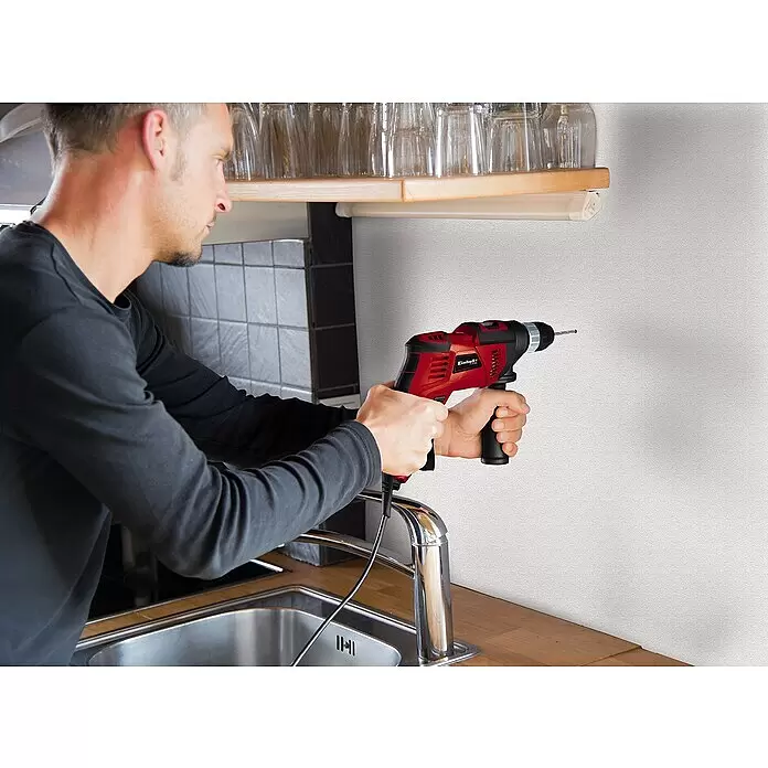 Einhell Taladro percutor TC-ID 1000 E Kit - 1.010 W, Portabrocas de sujeción rápida - Hombre sosteniendo un taladro Einhell rojo y negro, a punto de perforar una pared sobre un fregadero de cocina.