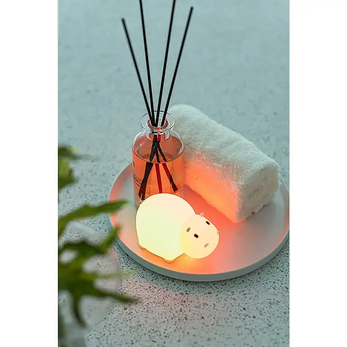 MegaLight LED-Nachtlicht SWIMMY HIPPO - L x B x H: 11 x 7 x 7 cm, Lichtfarbe: RGB, SWIMMY HIPPO - Stimmungsvolles Stillleben mit einem niedlichen, leuchtenden Hippo-Nachtlicht auf einem Teller neben einer Duftflasche und einem Handtuch.