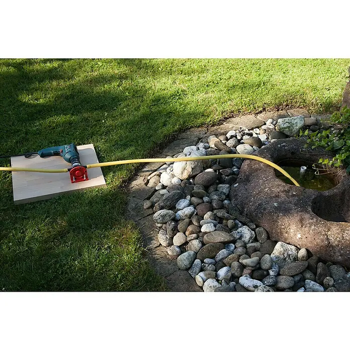 Wolfcraft Pumpe - 1.300 l/h, Förderhöhe: 15 m, Kunststoff - Ein Bohrer, der an eine Pumpe angeschlossen ist, befördert Wasser aus einem kleinen Teich in einem Garten. Die Pumpe steht auf einem Holzbrett.