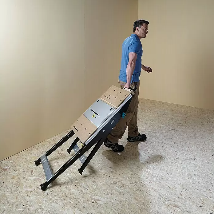 Wolfcraft Master Cut Werkbank 1500 - Arbeitshöhe: 865 mm, Belastbarkeit: 200 kg - Mann trägt eine klappbare Werkbank mit Holzarbeitsplatte, Metallelementen und schwarzen Beinen, was ihre Portabilität zeigt.