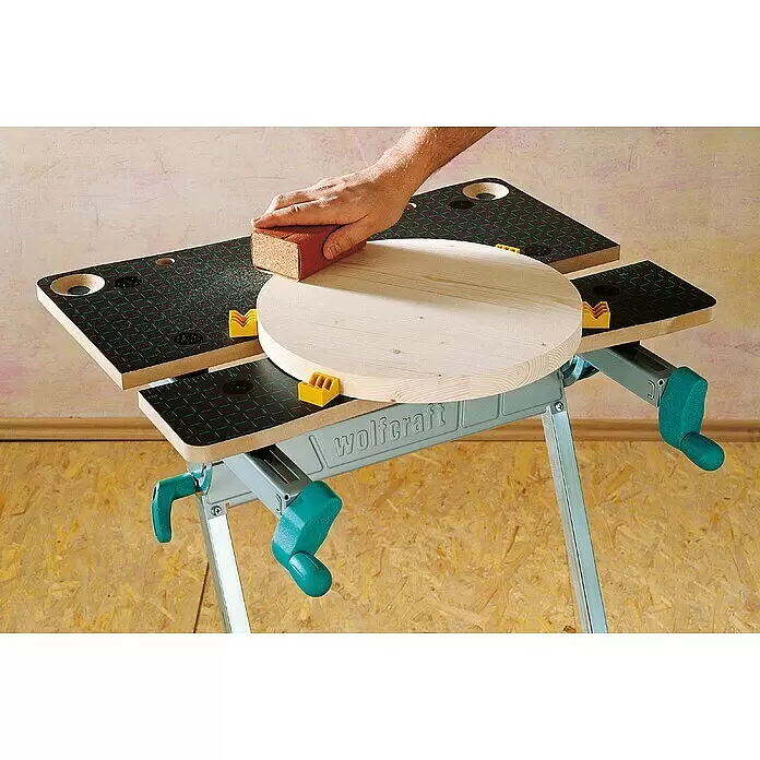 Wolfcraft Master Werkbank Master 600 - Arbeitshöhe: 950 mm, Belastbarkeit: 120 kg - Hand schleift eine runde Holzplatte auf einer grauen Wolfcraft Werkbank mit gelben Klemmen.