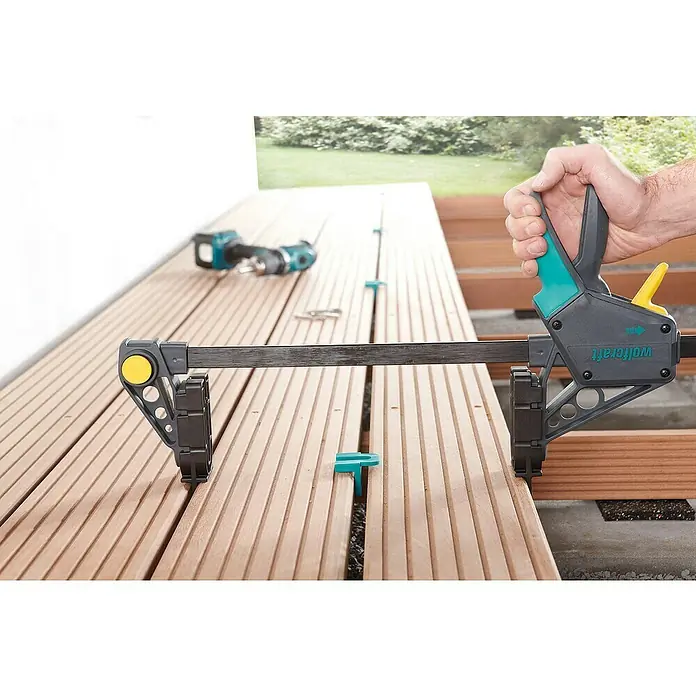 Wolfcraft Terrassenbau-Set - 11 -tlg. - Einhandzwinge von Wolfcraft im Einsatz, um Terrassendielen zu fixieren, während im Hintergrund Werkzeuge liegen.