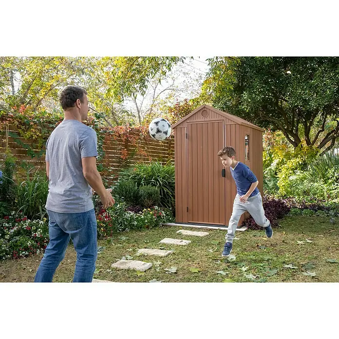 Keter Caseta de jardín Darwin 4x6 - Medidas ext. (An x Pr): 125,8 x 184,5 cm, Plástico, Marrón, 2,3 m² - Un padre y su hijo juegan al fútbol en el jardín, con un cobertizo de jardín marrón para guardar herramientas al fondo.
