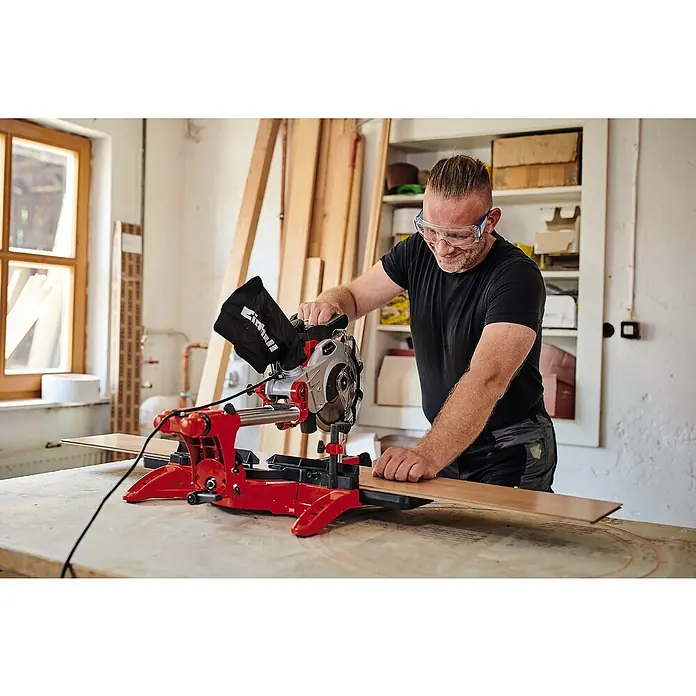 Einhell Kapp- & Gehrungssäge TC-SM 2131/1 Dual - 1.800 W, Durchmesser Sägeblatt: 210 mm, Leerlaufdrehzahl: 4.900 U/min - Ein Mann mit Schutzbrille bedient eine rote und silberne Einhell Kappsäge, um ein Holzbrett zuzuschneiden.