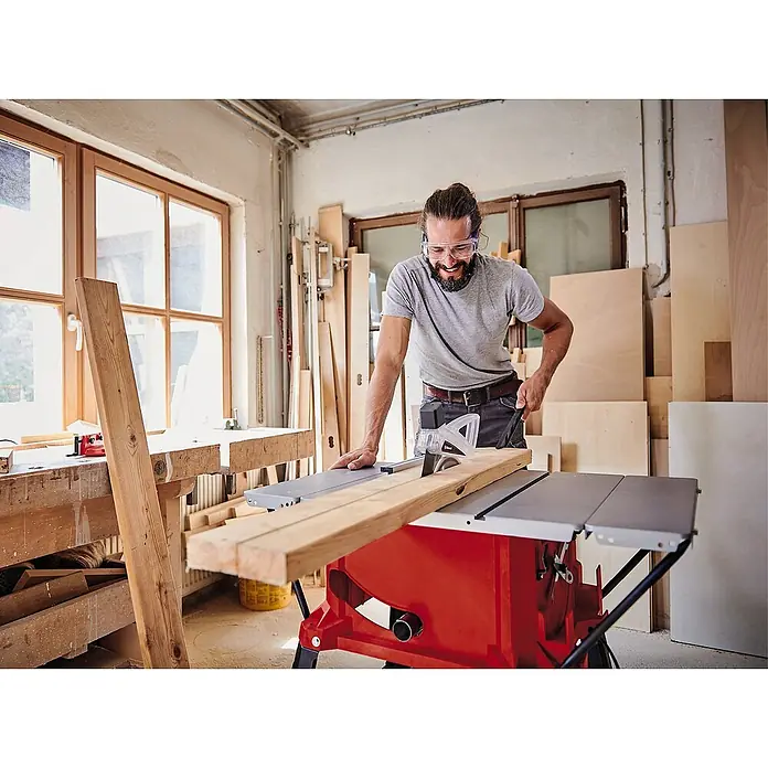 Einhell Sierra circular de mesa TC-TS 254 U - 1.800 W, Diámetro hoja de sierra: 254 mm, Profundidad de corte a 90 °: 80 mm - Un hombre usa una sierra de mesa Einhell roja para cortar con precisión una tabla de madera mientras usa gafas protectoras.