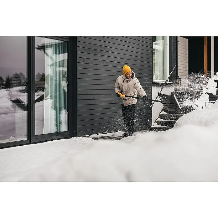 Fiskars Solid Schneeschieber - Breite: 54 cm, Langstiel - Mann schaufelt Schnee mit einer Schneeschaufel von Fiskars vor einem modernen Haus. Das Produkt ist ideal für die Schneebeseitigung rund ums Haus.