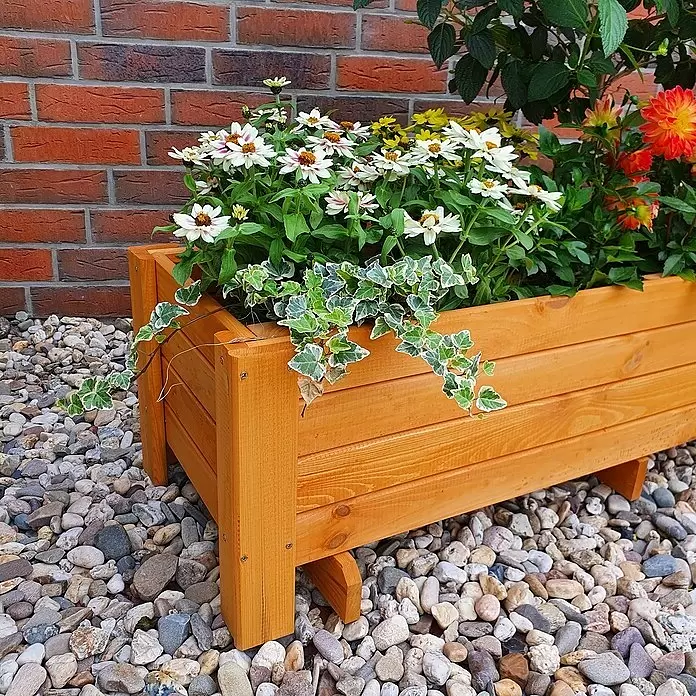 Promadino Pflanzkasten Romantica - Außenmaß (L x B x H): 118 x 38 x 34 cm, Holz, Honigbraun - Heller Holzpflanzkasten mit weißen, gelben und orangefarbenen Blumen sowie panaschiertem Efeu, auf Kies vor einer Ziegelwand.