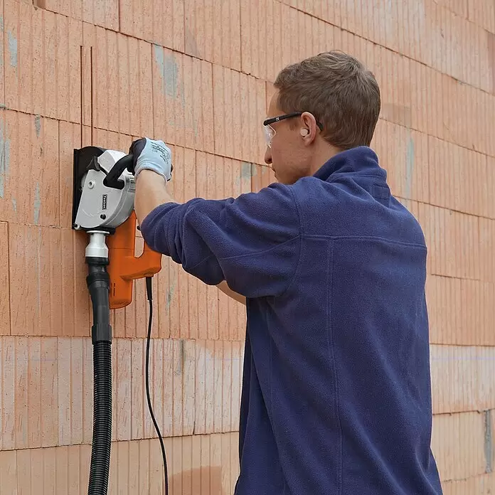 Toolson Mauernutfräse Pro-MF 1320 - 1.320 W, Durchmesser Scheibe: 125 mm - Mann nutzt eine silberne und orange Mauernutfräse mit Staubabsaugung, um Nuten in eine Ziegelwand zu schneiden.