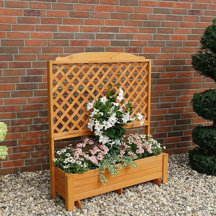 Promadino Pflanzkasten Romantica - Außenmaß (L x B x H): 120 x 38 x 140 cm, Holz, Honigbraun - Holzpflanzgefäß mit Gitterwerk und Blumen. Der Blumenkasten mit Rankgitter ist bepflanzt mit rosa und weißen Blumen, vor einer Backsteinmauer.