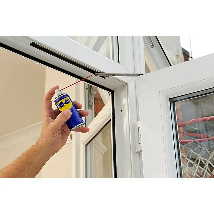 WD-40 Aceite para herrajes multiuso - 100 ml - Una mano rociando producto multiusos WD-40 en un marco de ventana para lubricar y mejorar la funcionalidad.