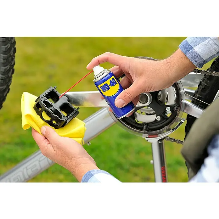 WD-40 Aceite para herrajes multiuso - 100 ml - Un hombre limpiando un pedal de bicicleta con un paño amarillo mientras aplica WD-40 con una pajita roja para lubricarlo.