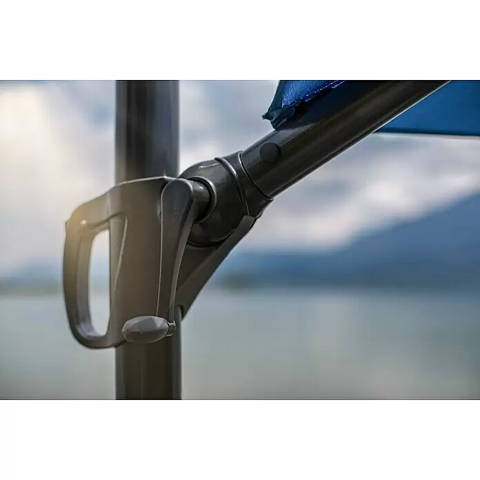 Sunfun Ampelschirm rund Florenz - Blau, Durchmesser Bespannung: 300 cm - Dunkelgrauer Sonnenschirm-Kurbelmechanismus und Arm, mit blauem Stoffteil, vor unscharfem Outdoor-Hintergrund aus Himmel und Wasser.
