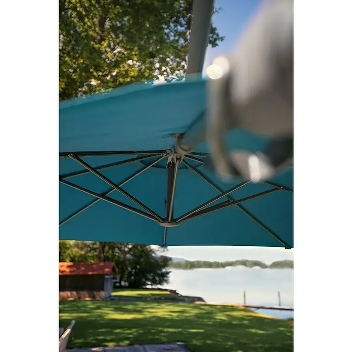 Sunfun Schirmbezug Capri - Petrol, Passend für: Sunfun Ampelschirmgestell Capri Ø 300 cm - Blick unter einen blauen Sonnenschirm auf einen See. Der Schirm spendet Schatten und bietet Schutz vor der Sonne.
