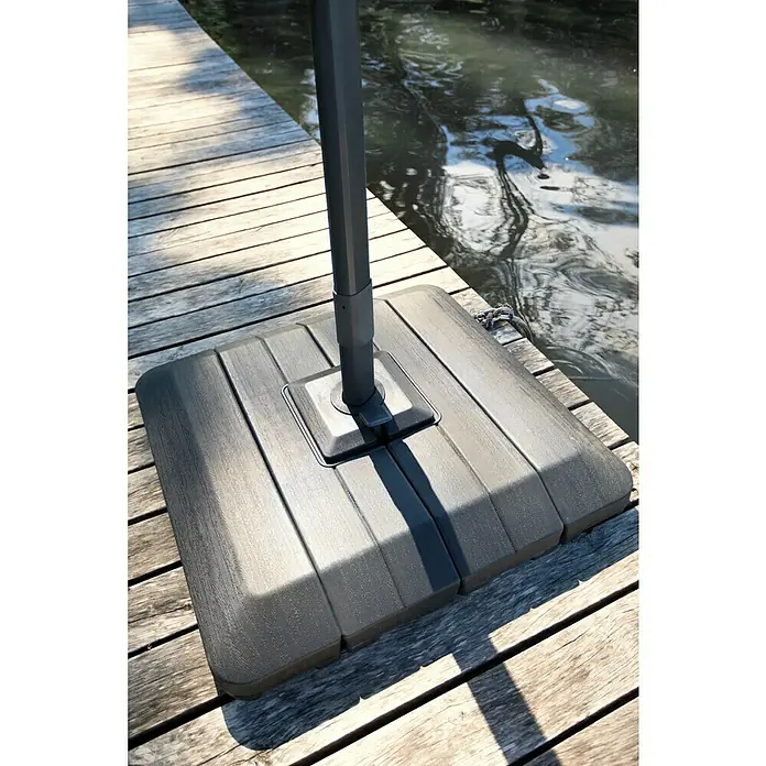 Sunfun Ampelschirm Capri - Anthrazit, Bespannungsmaß (L x B): 300 x 350 cm - Nahaufnahme eines grauen, quadratischen Sonnenschirmständers aus Kunststoff mit Holzstruktur auf einem hölzernen Steg am Wasser.