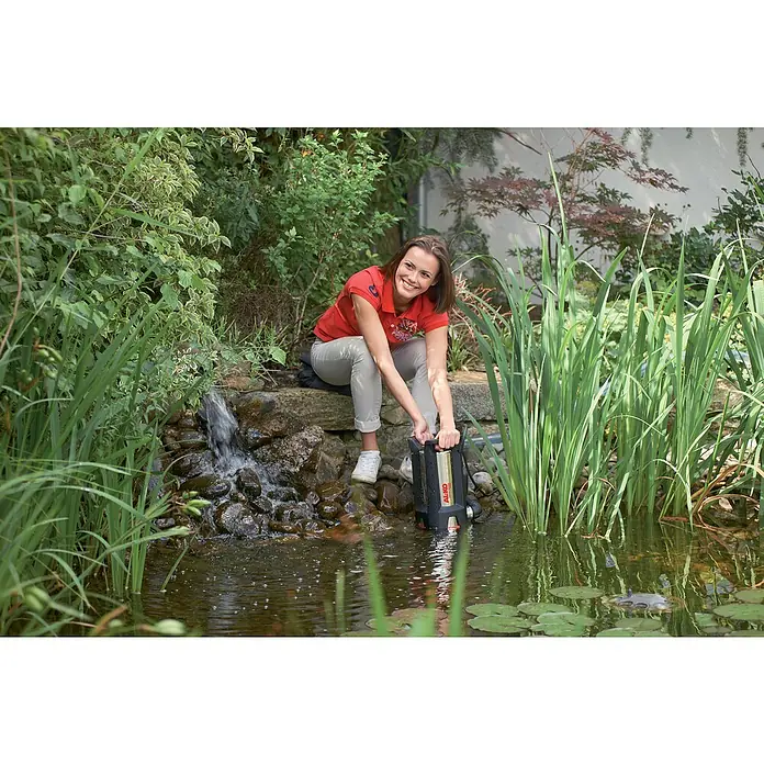 AL-KO Comfort Schmutzwasserpumpe Drain 10000 Inox - 750 W, Max. Fördermenge: 10.000 l/h, Eintauchtiefe: 7 m - Frau hockt an einem Teich und bedient eine schwarze AL-KO-Teichpumpe, umgeben von üppigem Grün.