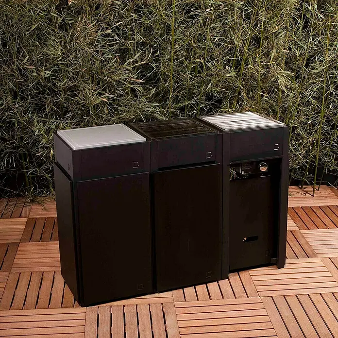 oneQ Blende - Schwarz, Passend für: oneQ Basismodul - Drei stilvolle Outdoor-Küchenmodule aus dunklem Material stehen auf einer Holzterrasse vor einem grünen Hintergrund.