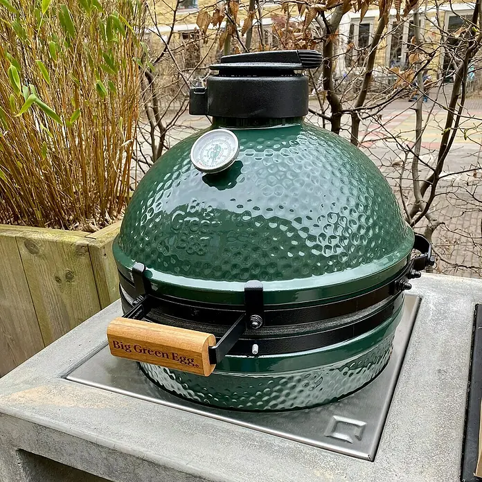 oneQ Grilleinsatz Kamado Grill - Passend für: Kamado Joe Holzkohlegrill Joe Junior / oneQ Basismodul / oneQ Einbausockel, L x B: 40,7 x 40,7 cm, Edelstahl - Ein Big Green Egg Keramikgrill in Grün, platziert auf einem Betontisch im Freien, umgeben von Pflanzen.