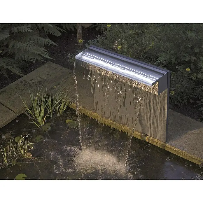 Ubbink Einbau-Wasserfall Niagara - 12,5 x 30 x 10 cm, Edelstahl, LED - Nahaufnahme eines modernen Wasserfall-Gartenbrunnens mit LED-Beleuchtung, der in einen Teich mündet, umgeben von üppigem Grün.