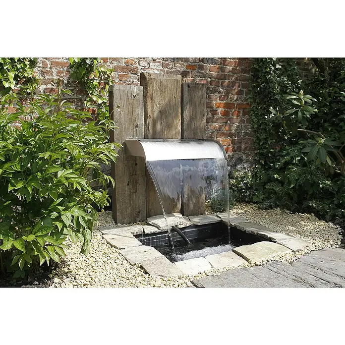 Ubbink Einbau-Wasserfall Nevada - 33 x 90 x 13 cm, Edelstahl, LED - Wasserspiel mit Wasserfall aus Edelstahl, umgeben von Steinen und Pflanzen, eingebettet in eine grüne Gartenlandschaft vor einer Backsteinmauer.