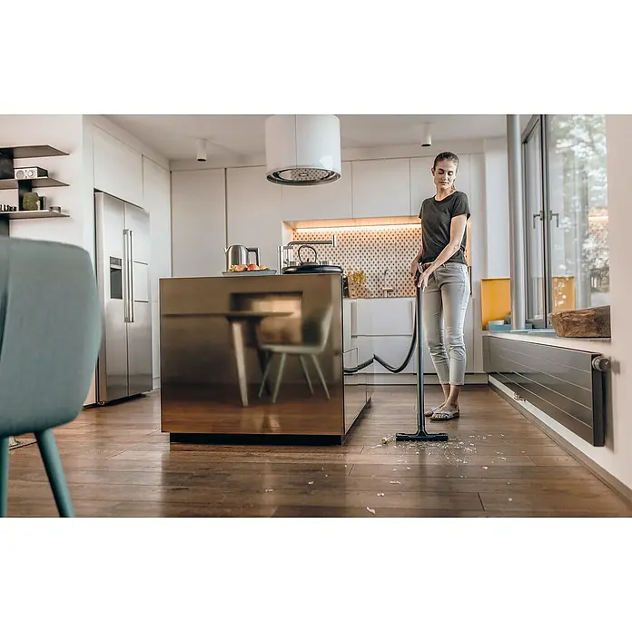 Kärcher Aspirador en seco y húmedo eléctrico KWD1 - 1.000 W, Volumen de recipiente: 12 l, Con bolsa - Una mujer utiliza una aspiradora Kärcher para limpiar vidrios rotos en una cocina moderna, enfatizando la eficiencia de limpieza de la aspiradora.
