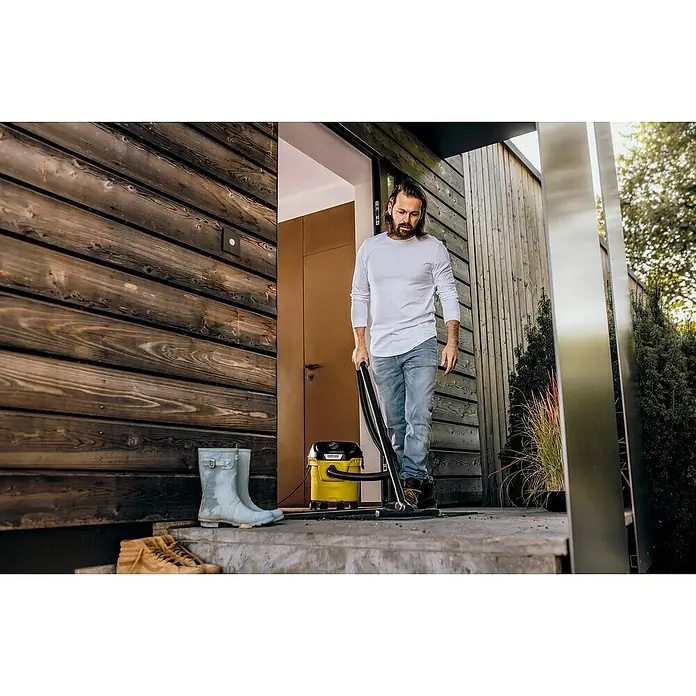 Kärcher Aspirador en seco y húmedo eléctrico KWD1 - 1.000 W, Volumen de recipiente: 12 l, Con bolsa - Un hombre está usando una aspiradora multiusos Kärcher para limpiar un porche. La aspiradora es amarilla y negra, y el hombre lleva vaqueros y una camisa blanca.