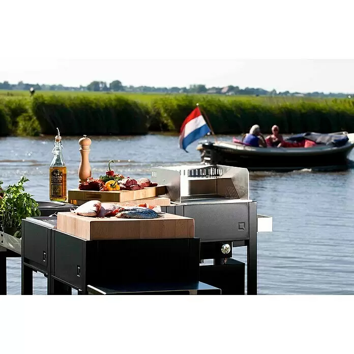 oneQ Basismodul - L x B x H: 44 x 44 x 92 cm, Schwarz, Metall - Schwarzer Outdoor-Grill mit integrierten Holzschneidebrettern, beladen mit Fisch, Fleisch und Gemüse, an einem malerischen Fluss.