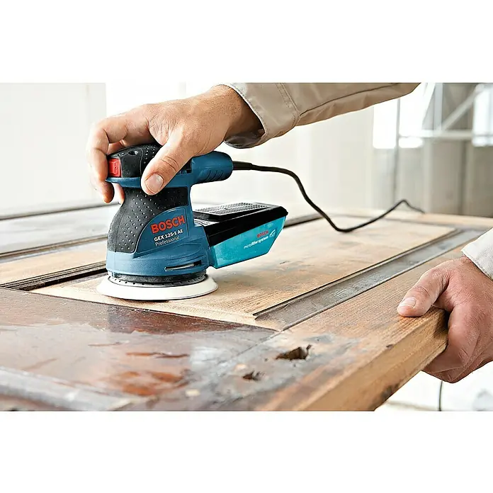 Bosch Professional Elektro-Exzenterschleifer GEX 125-1 AE - 250 W, Durchmesser Schleifteller: 125 mm, Leerlaufdrehzahl: 7.500 U/min - 12.000 U/min - Nahaufnahme des Bosch GEX 125-1 AE Exzenterschleifers in Aktion, der eine Holzoberfläche glättet.