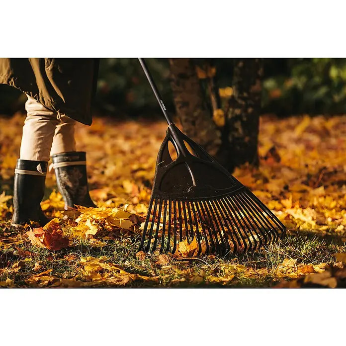 Fiskars Solid Laubbesen XL - Arbeitsbreite: 65 cm, Anzahl Zinken: 27, Langstiel - Nahaufnahme eines Laubbesens der Marke Fiskars, der auf einer mit Herbstlaub bedeckten Rasenfläche liegt, während eine Person im Hintergrund steht.
