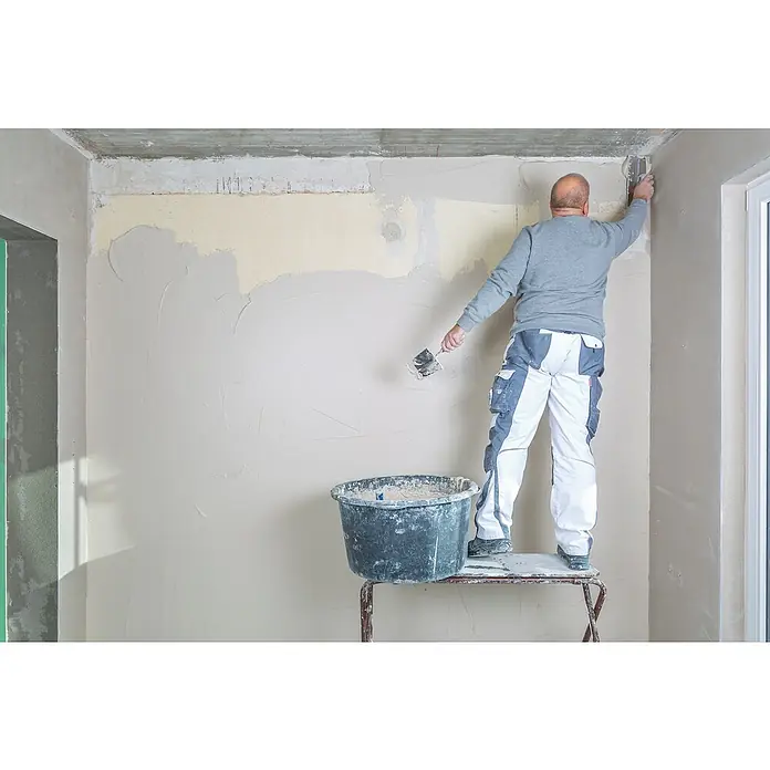 Knauf Rotband Flächenspachtel - 5 kg - Ein Bauarbeiter auf einer Leiter spachtelt mit Spachtelmasse eine Wand, um eine glatte Oberfläche zu erzielen, ideal für Malerarbeiten.