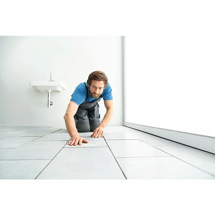 Knauf Flexfuge Universal - Manhattan, 10 kg - Ein Handwerker kniet auf dem Boden und verlegt Fliesen in einem Badezimmer, um das neue Badezimmer zu installieren.