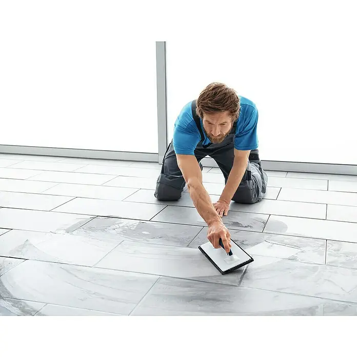 Knauf Flexfuge Universal - Manhattan, 10 kg - Fliesenleger kniend, mit Werkzeug in der Hand, reinigt er die neu verlegten Bodenfliesen, um ein sauberes Finish zu gewährleisten.